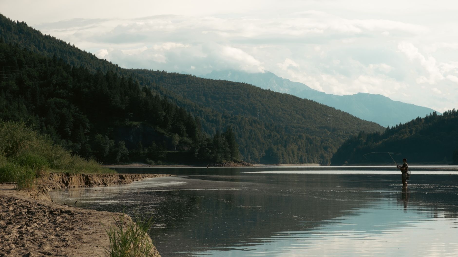 Fisker står i et stille skogsvann omgitt av skogkledde fjell